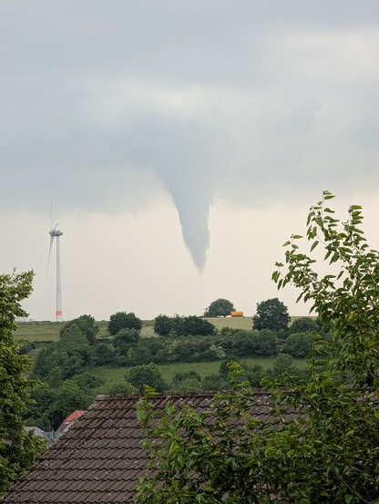 Grauer Himmel aus dem eine Wolke wie ein Eiszapfen nach unten herausschaut. Daneben ein Windrad. 