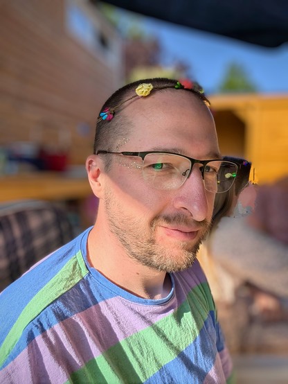 Person lächelt seitlich in die Kamera. Auf ihrem Kopf ein Blütenkranz aus Plastik. Neben ihren Augen ist ein Kringel aus Glitzer auf der Haut. Sie trägt ein buntes, gestreiftes Shirt und eine Brille. Die Person sitzt im Freien unter einem Schirm. Im nachträglich verschwommenen gemachten Hintergrund ist ein Holzhaus und zwei Personen zu erkennen.