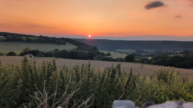 Zeitraffer eines Sonnenuntergangs. Oberstes Drittel des Videos zeigt hell- bis dunkelorangen Himmel. Darunter ein Streifen dunkelgrüner Wald. Im untersten Drittel hellgrüne Wiesen und im Vordergrund dunkelgrüne Pflanzen und verschwommen Steine. 