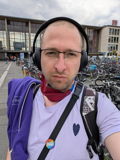Person mit kur rasierten Haaren, Brille, FFP-Maske um den Hals gehängt und Kopfhöreren auf dem Kopf steht vor dem Bahnhof in Heidelberg. Im Hintergrund sehr viele Fahrräder. Die Person trägt einen Rucksack und eine Schultertasche. Auf dem schwarzen Träger der Schultertasche ist ein regenbogenfarbener Actually Autistic Pin. Das helllila T-Shirt hat ein dunkellila Herz aufgestickt. Über der Schulter hängt ein dunkellila Hoodie, auf dem man das pretix Logo erkennen kann. 