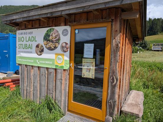 Eine etwa drei Mal zwei Meter große Holzhütten auf einem Feld. Vorne ist eine Glastüre, wie sie oft bei Terrassen verwendet wird und daneben ein Plakat: Team Biohof. Bio Ladl Stubai.

Rechts neben der Hütte ist ein Brett zum Sitzen. 