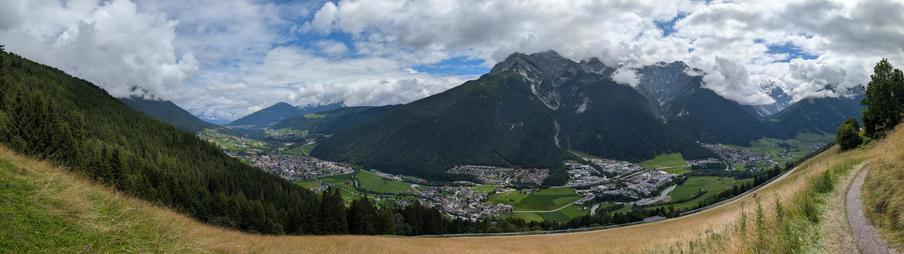 Panorama eines Tales von einer Talseite. Blauer Himmel mit vielen weißen Wolken. Im Tal mehrere Ortschaften. Am Rand des Panoramas ein Weg.