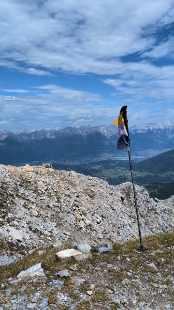 Selbstgemalte Nichtbinär-Flagge weht an einem Wanderstock. Im Hintergrund Blick auf das Inntal mit der Stadt Innsbruck und dahinter die Gebirgskette Nordkette. Blauer Himmel mit einigen weißen Wolken. 