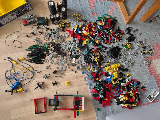 Fußboden mit über dreißig Häufchen an Lego Technik Teilen. Links oben ist das Eck einer Lego Technik Verpackung. Darunter zwei Steuereinheiten und ein Motor. Daneben ein paar Kabel und mehrere Reifenstapel. Links in der Mitte mehrere Pumpen und hydraulische Elemente, die mit dünnen Schläuchen verbunden sind. Häufchen mit Zahnrädern, Verbindungsstangen, Steckverbindern, Platten, längliche Teile, Teile mit schrägen Kanten, hohe Teile mit und ohne Löchern. 