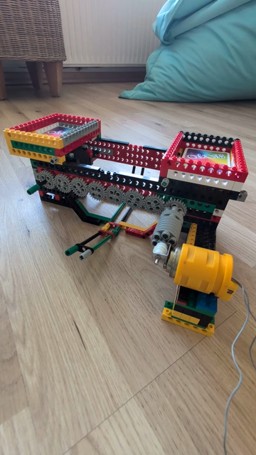 Die Kartenmischmaschine aus Lego Technik in Betrieb. Links und rechts sind zwei Säulen, auf denen die rechteckigen Behälter für die Karten befestigt sind. Oben sind die Behälter miteinander verbunden und an der Außenseite acht Zahnräder, die die beiden Gummiräder unter den Kartenbehältern mit dem Motor verbinden, der sich auf der rechten Seite der Konstruktion befindet. Die Karten fallen erst langsam und dann immer schneller in den Behälter der lose in der Mitte unter dem Gerät steht und nur au…