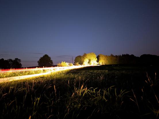 Foto mit langer Belichtungszeit. Obere Hälfte Nachthimmel mit einigen Sternen, aber keinen Sternschnuppen. Untere Hälfte Wiese mit ein paar Bäumen am Horizont. Von der linken Seite zieht sich ein heller Lichtstreifen leicht schräg durch das Bild. Das Auto selbst ist aufgrund der langen Belichtung nicht zu sehen. Teilweise sind auch Gräser und Bäume angeleuchtet.