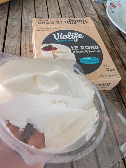 Im Vordergrund eine einheitlich weiße Masse in einer runden Plastikverpackung. Im Hintergrund die Kartonumpackung in Holzoptik. Mittig darauf ein Foto der veganen Lebensmittelzubereitung auf schwarzen Hintergrund mit einem Klecks Preiselbeermarmelade und drei Rosmarinstängel.