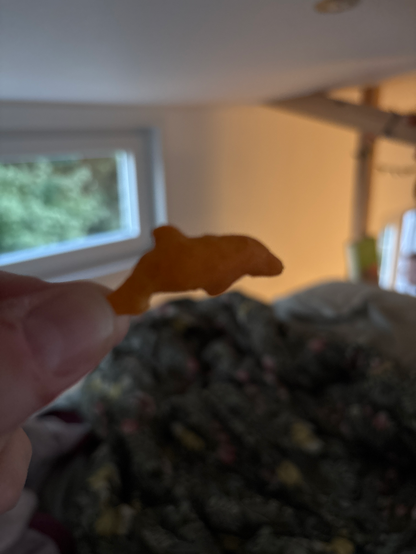 Ein Kartoffelchip-Delphin, der mit zwei Fingern vor die Kamera gehalten wird. Im Hintergrund Bettzeug.
