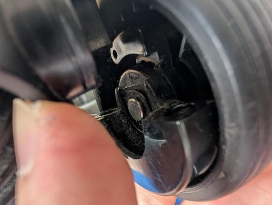 Nahaufnahme einer Radaufhängung einer Staubsaugerbürste. Ein Finger drückt ein Plastikteil zur Seite. Daneben ist das Gummirad mit einem Metallbolzen in der Aufhängung. 