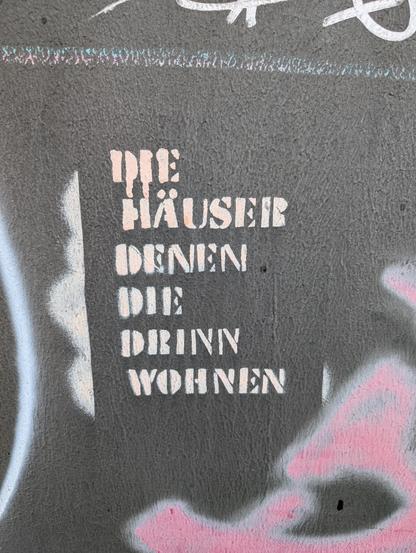Graffiti. Weißer Stencil auf dunkelgrauem Hintergrund mit dem Text:
Die Häuser denen die drinn wohnen