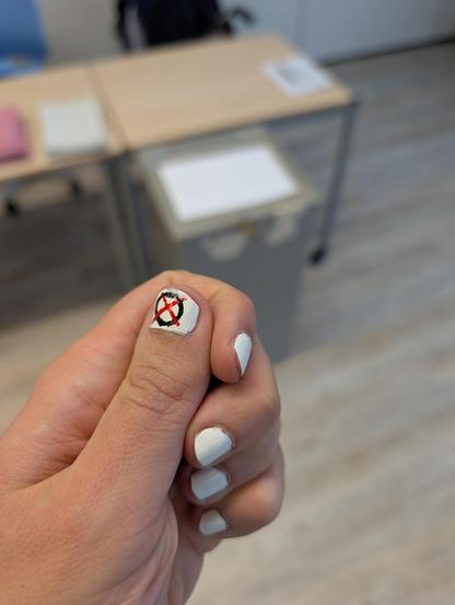 Im Vordergrund eine Hand mit weiß lackierten Fingernägeln. Auf dem Daumen ist ein schwarzer Kreis mit einem roten X aufgemalt. Die Striche des X gehen leicht über den Kreis hinaus. Im Hintergrund verschwommen eine Wahlurne und Tische dahinter, auf denen Zettel liegen. 