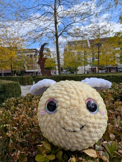 Häkelbiene auf einem Busch. Im Hintergrund eine Metallskulptur, die ein Gesicht darstellt.