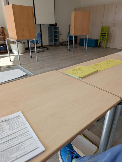 Im Vordergrund ein Stapel mit Wahlzettel auf einem Tisch. Links eine Wahlurne mit einem Zettel abgedeckt. Im Hintergrund zwei Tische mit Holzabschirmungen als Wahlkabinen. Daneben eine Tafel und ein Stapel hellgrüner Stühle.