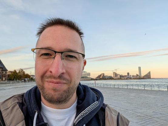 Luca, eine Person Mitte dreißig, steht leicht lächelnd vor dem Meer. Im Hintergrund unterschiedliche Hochhäuser auf einer Landzunge.