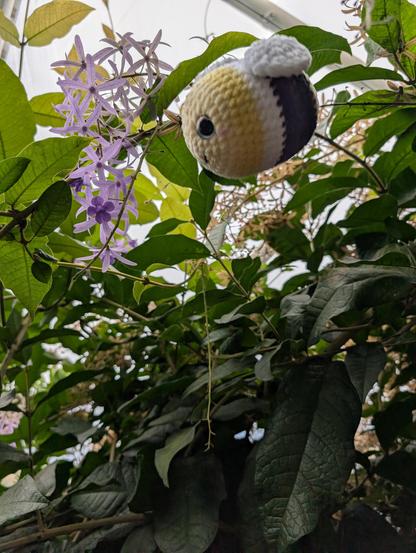 Crochet bee in non binary colors flies towards a violett flower. Green leafs in the background.