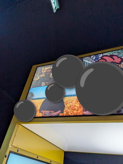 Photo of a screen with multiple photos at an interactive station in the museum. The crochet bee sits on the shoulder of a person wearing a pretix shirt. The people in the other photos are hidden behind black emoji circles. 