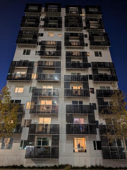 Hochhaus in der Nacht. Um die Balkone sind jeweils zehn bifaziale Photovoltaikmodule in einem Rechteck angeordnet. Dazwischen sind teilweise Löcher, wo keine Balkone sind. 