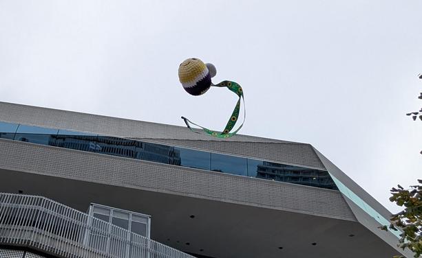 Crochet bee with a green lanyard attached to it. On the lanyard are sunflowers. In the background is a futuristic building.