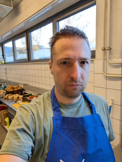 Person Mitte Dreißig mit zerzausten Haaren und müden Blick schaut in die Kamera. Im Hintergrund geschnittene Brötchen und Belag auf einer Edelstahlarbeitsplatte.