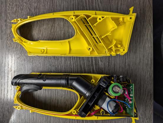 Fenstersauger in zwei Teile zerlegt. Oben eine Hälfte aus gelben Plastik. Unten die zweite Hälfte, in der noch die restlichen Teile sind. Zwei schwarze Rohrstücke. Das untere mit einem Knick. Dann kommt das Schaufelrad oder wie man das nennt, das die Luft nach außen drängt. Daran der Motor. Ganz unten eine Platine und eine 18650VT Lithium-Ionen-Batterie.