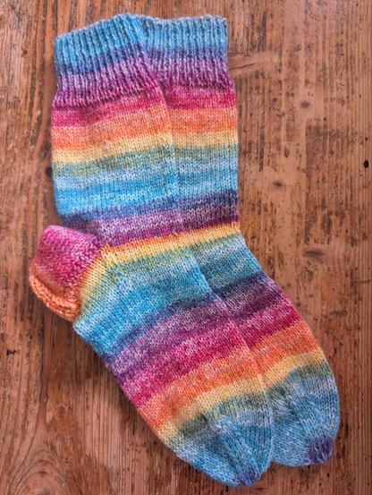 Wollsocken in Regenbogenfarben gestreift. Ferse und Zehenbereich ist verstärkt. Die Socken liegen auf einer Holzfläche. 