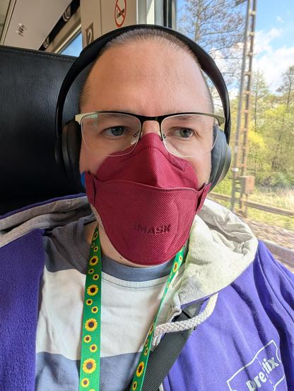 Person mit kurz rasierten Haaren sitzt in einem Zug. Sie trägt Kopfhörer, eine rote FFP2-Maske, ein grünes Autismuslanyard mit Sonnenblumen und eine lila pretix-Jacke. Der Blick geht leicht an der Kamera vorbei.