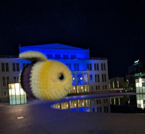 Gehäkelte Biene in den Farben der Non Binary Flagge in der Luft vor einem blau beleuchteten Gebäude. 