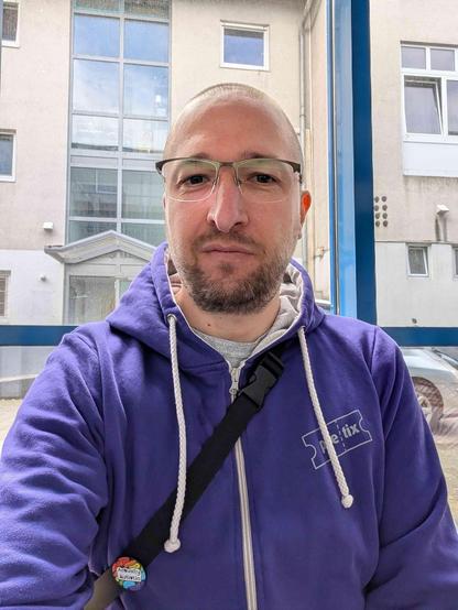 Person Mitte Dreißig mit lila Pullover und Brille sitzt vor einer Glasscheibe.
