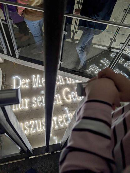 Ein Metallsteg mit Personen (nur Unterkörper) der zick zack mehrere Meter über dem Boden ist. Auf dem Boden stehen in weißer Leuchtstoffröhrenschrift etwas, das durch den Steg so verdeckt wird, dass es nicht vollständig lesbar ist. Ein Teil des Textes spiegelt sich im Glasgeländer des Stegs.